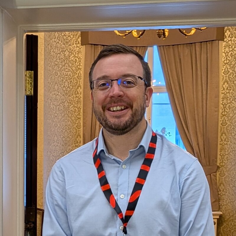 Oliver (Ollie) Cronk smiling, wearing a light blue shirt and a speaker lanyard at a conference